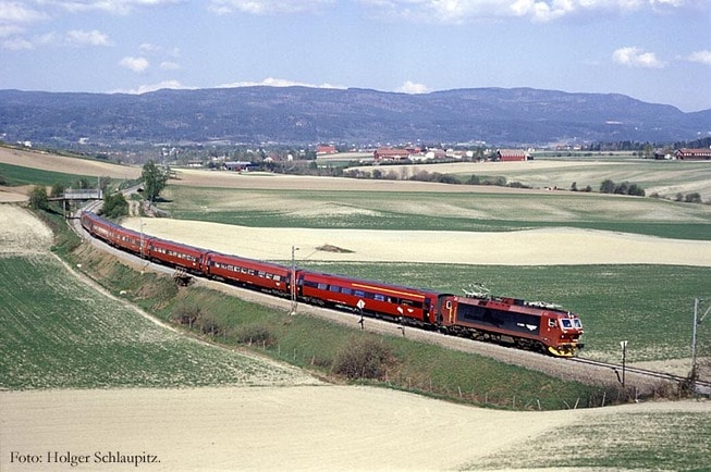 Et rødt tog kjører gjennom et nokså flatt jordbrukslandskap