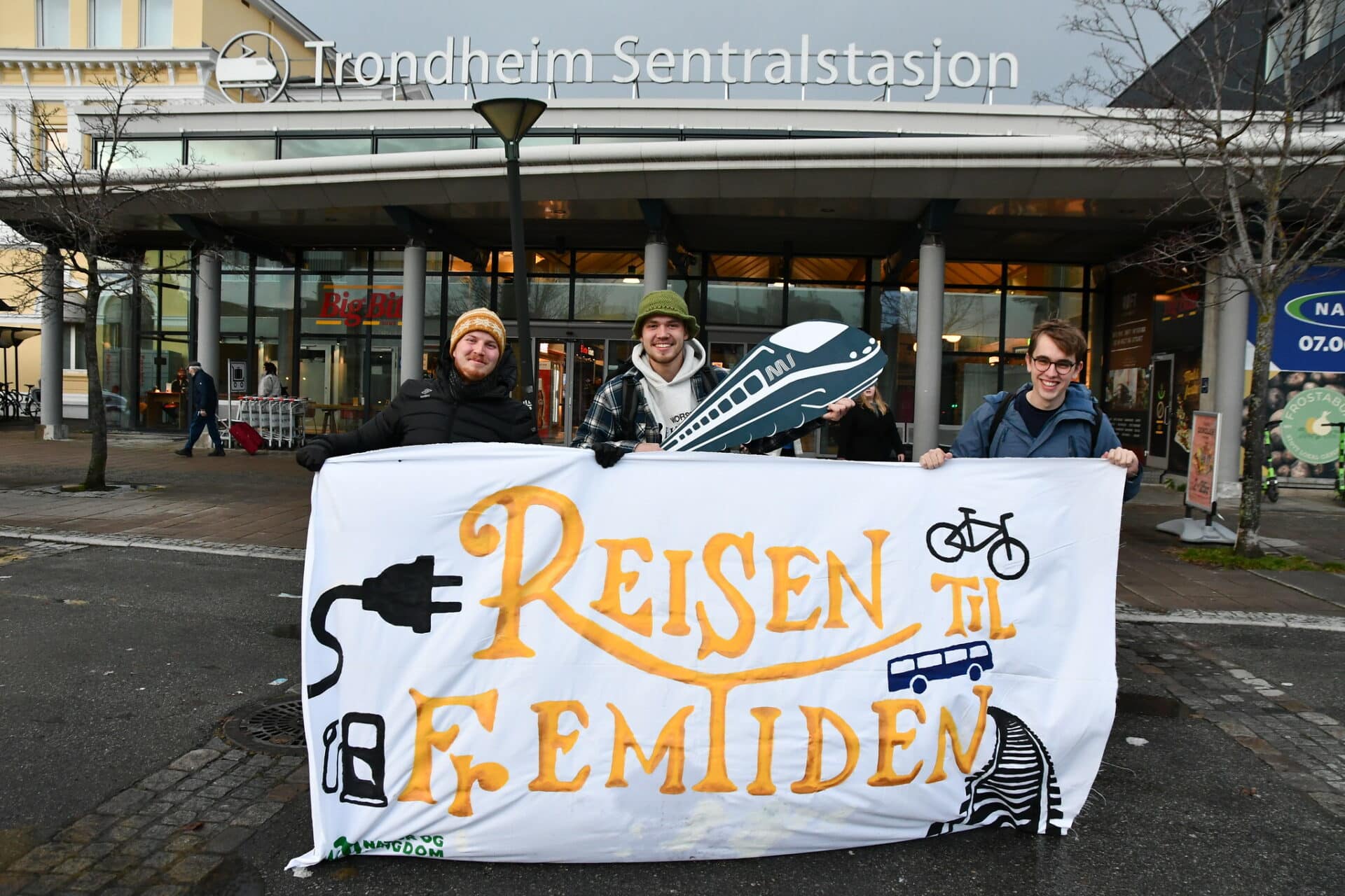 Tre ungdommer står foran Trondheim sentralstasjon med et banner hvor det står "Reisen til fremtiden" med gul skrift. På banneret er det malt en sykkel, et støpsel og et sett togskinner.