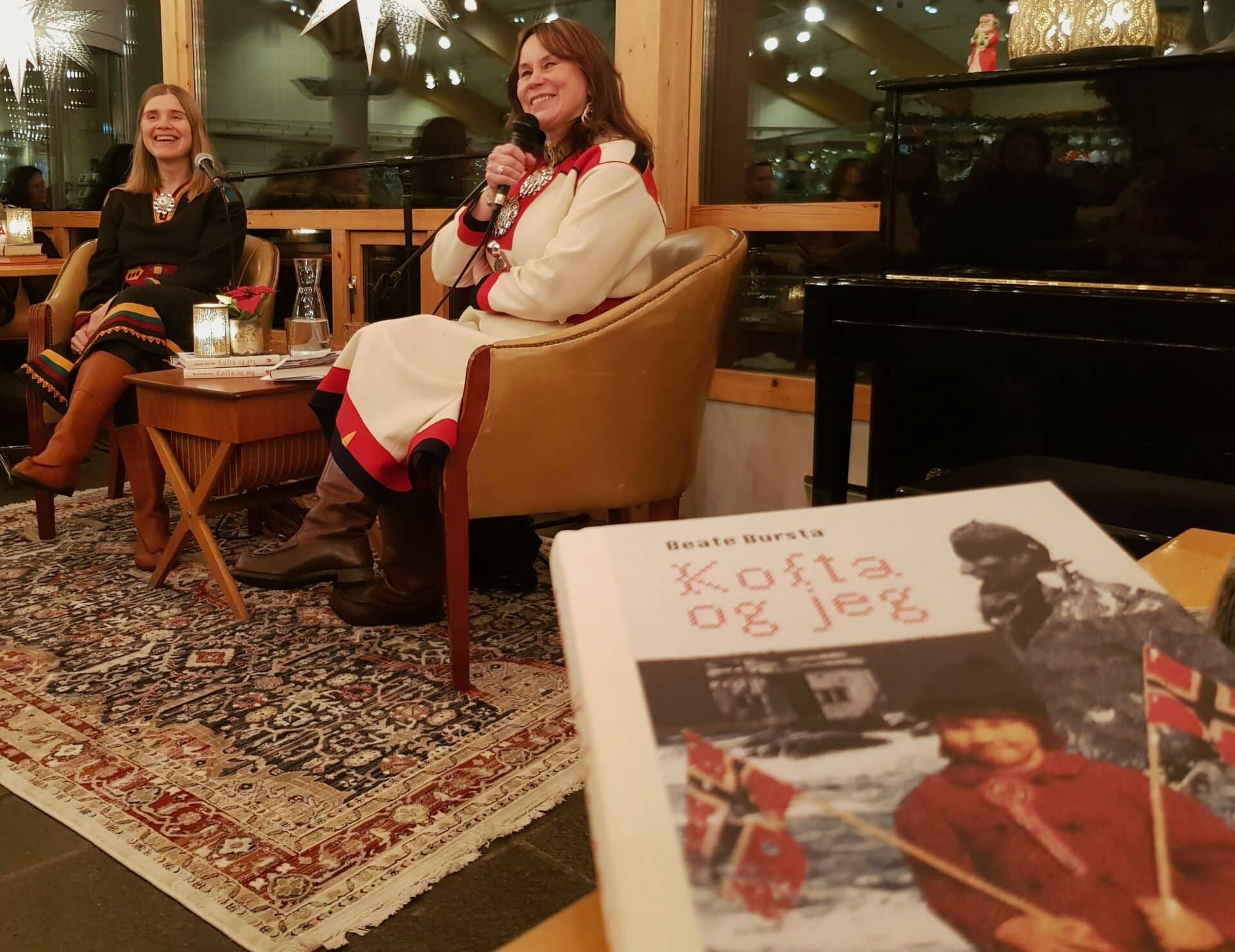 To kvinner i kofte prater sammen, sittende i hver sin lenestol. I forgrunnen ser vi omslaget på ei bok: Kofta og jeg.