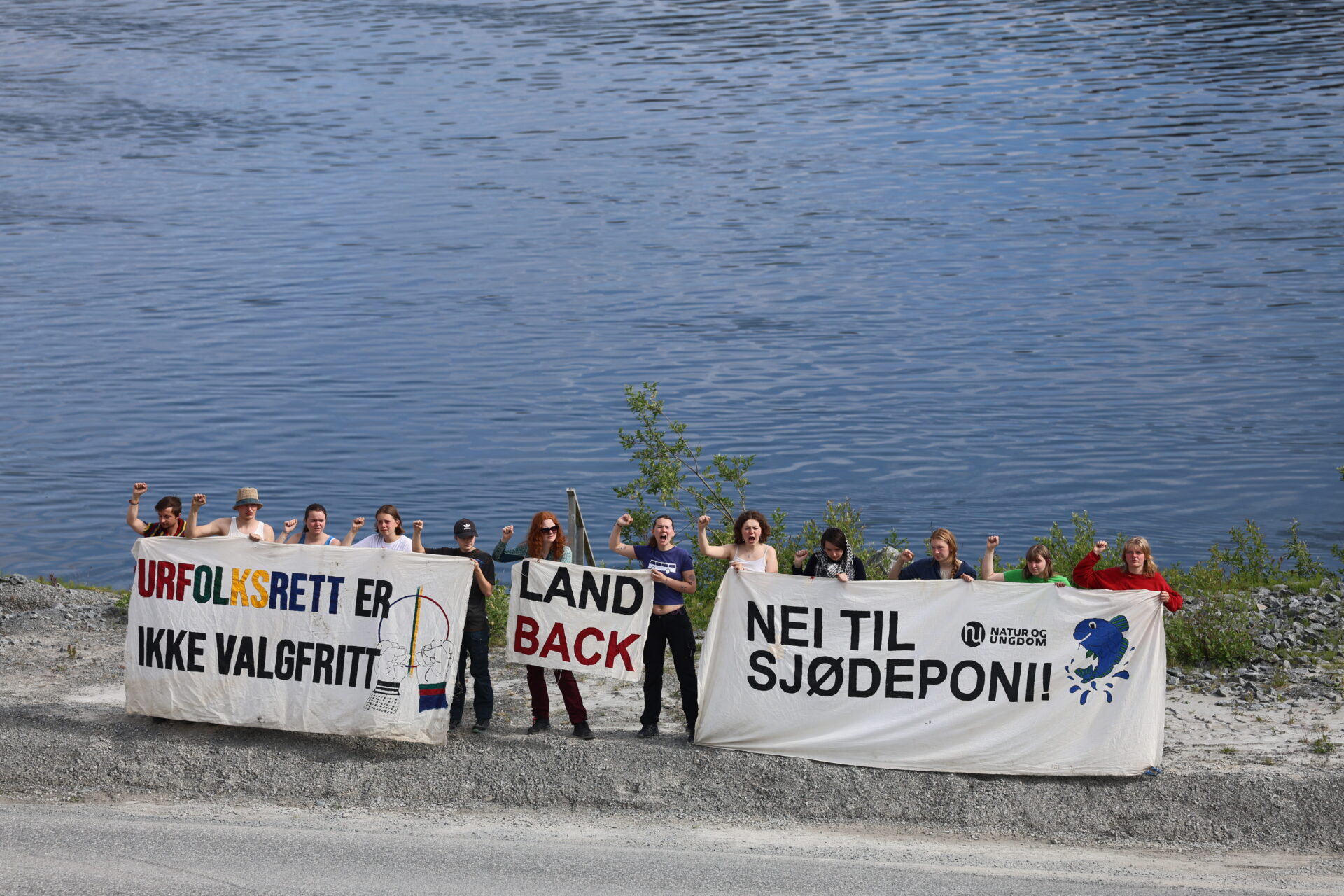 Demonstrasjon ved Repparfjorden i forbindelse med arbeid ved Nussirgruven sommeren 2025. Unge aktivister holder bannere med slagordene "Urfolksrett er ikke valgfritt", "Land back" og "Nei til sjødeponi".