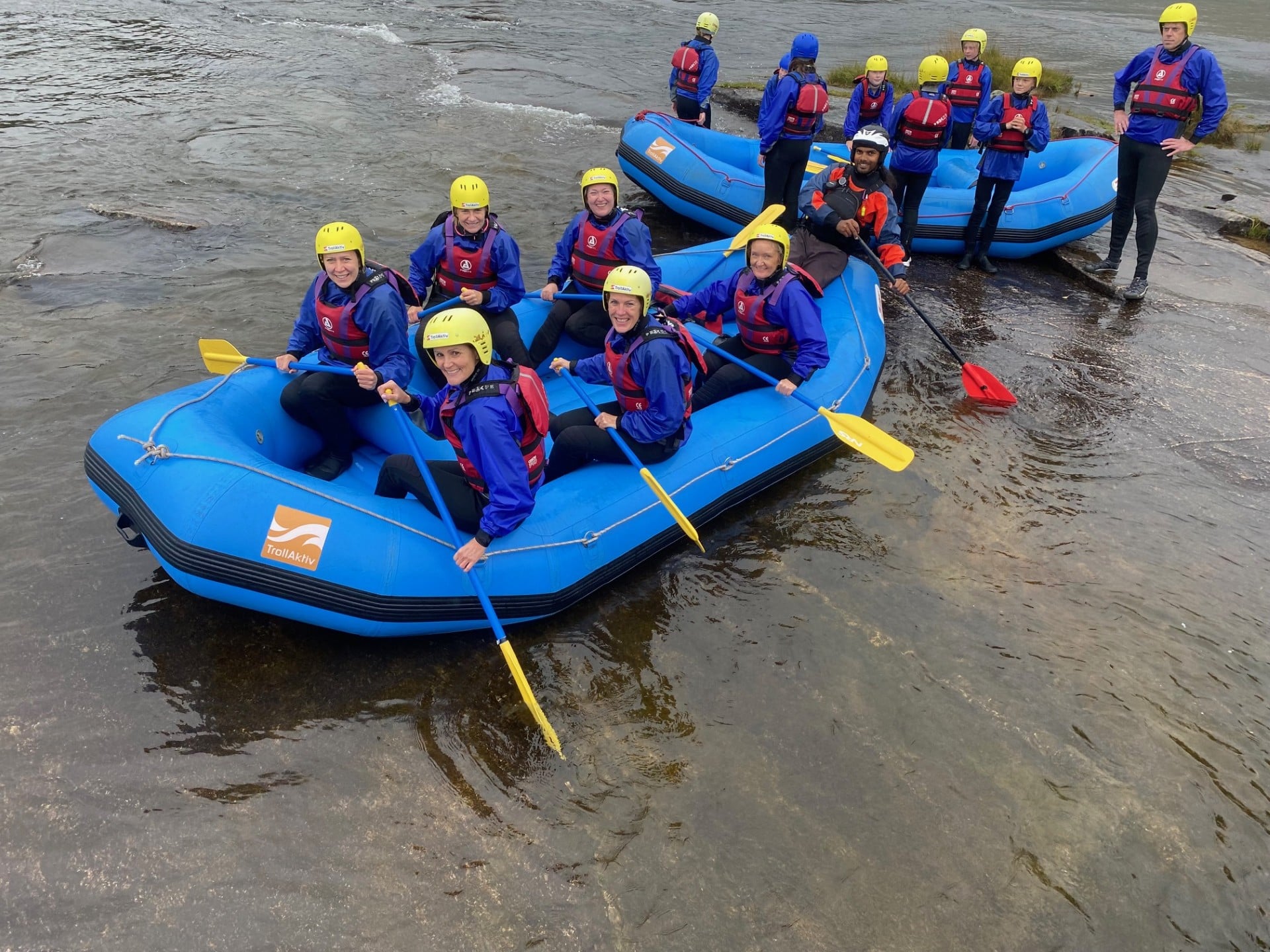 En gruppe mennesker sitter i blå rafting-flåter inntil elvebredden. Noen står utenfor båtene. 